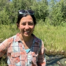 woman in an orange and while plaid shirt stands in front of a grassy field with trees