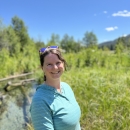 a woman in a blue long sleeve shirt and sunglasses on her head smiles with green field and river in the background