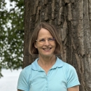 a white woman with blond hair and blue eyes, wearing glasses and a light blue polo shirt, in front of a tree, with a lake in the background