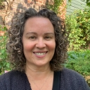 A woman wearing a black V-neck shirt with curly dark, shoulder length hair is smiling at the camera