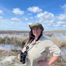 A U.S. Fish and Wildlife Service employee wearing a hat and binoculars stands near a wetland under a partially cloudy sky.