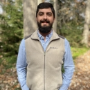 Alex stands smiling wiht hands in his pockets. He is wearing a tan fleece vest and a light blue button down shirt