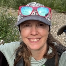 A person with long hair wearing sunglasses, a hat, and a personal flotation device vest smiles in the outdoors.