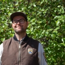 man in brown vest smiles in front of green trees