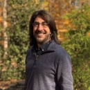 A person standing against a background of trees wearing glasses, and smiling. 