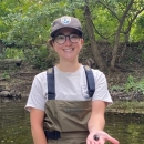a woman stands in a stream with trees in the background.