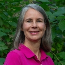 Woman head and shoulders, smiling and wearing a bright pink collared shirt. Green foliage in the background.