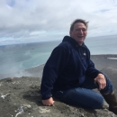 Man in sweatshirt sits on brown island with pale ocean and cloudy sky behind him.