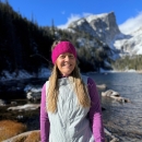 A woman with long light brown hair stands surrounded by mountains