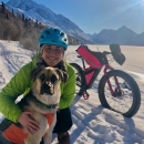 A picture of a woman next to a dog with a bicycle and snowy mountain landscape in the background