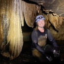 A person kneeling in a cave next to drapery formations. 