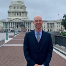 Ryan in a blue suit and tie stands smiling with hands clasped in front of the House of Representatives