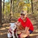 Shannon kneels and smiles with her arms wrapped around a white and brown spotted pitbull. She wears black running shorts, red running shoes, a red t-shirt, and a tan baseball cap