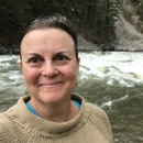 Yvonne smiles in a tan sweater with a blue shirt visible underneath in front of a running body of water