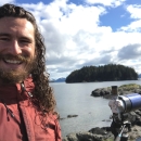 A man standing next to a spotting scope with a rocky shoreline and island in the background