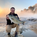 Man kneels at water's edge holding a large fish." class=" lazyloaded">