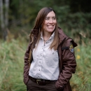 A woman with long brown hair wears a Refuges uniform brown jacket, tan shirt, and brown pants and stands outside.