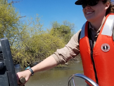 A woman drives a boat wearing a life vest and Fish and Wildlife Service hat