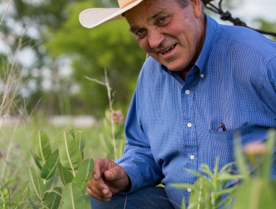 Bill Sproul, a rancher wearing a button-up blue shirt and ivory-colored cowboy hat, crouches in grass with his horse visible in the background