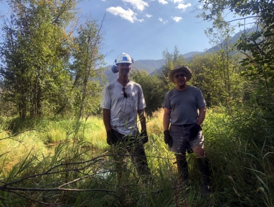 A tall man in hardhat, dirty t-shirt, and ear protection stands next to a shorter man in straw hat, shorts, and t-shirt, both wearing work gloves, in a field.