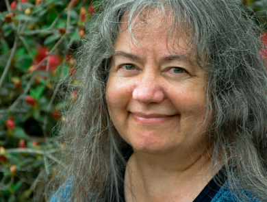 A smiling woman with long, gray hair and blue shirt and sweater stands in front of a green-leaved shrub with orange flowers
