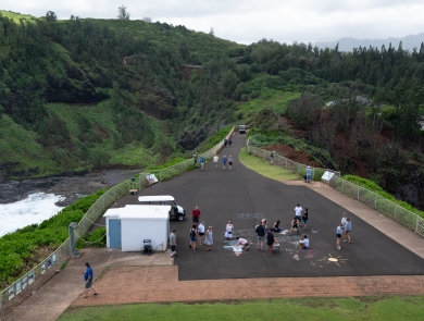 Visitors on a large black walkway at top a bluff are walking and making chalk art. The walk way is surrounded by lush green vegetation and an ocean cove can be seen on the left side. 