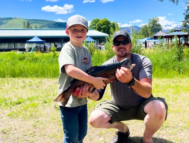 A young boy and his dad show off a caught large trout in front of the fishing pond