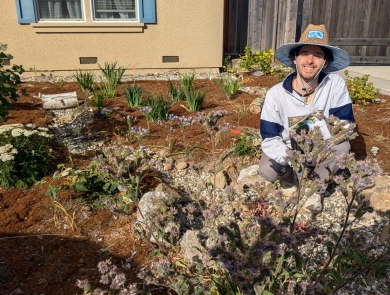 a man wearing a wide brimmed hat squats among plants in a front yard.