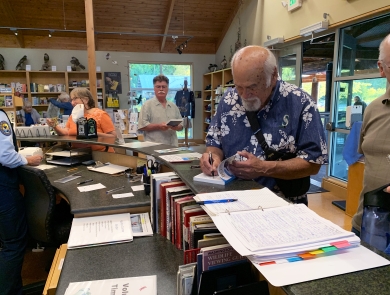 A man with white hair and beard and wearing a Hawai'ian shirt signs a book at the counter of a visitor center store.