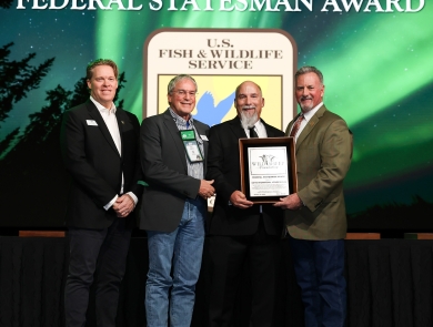 Four people stand onstage and in front of a screen that has the USFWS logo with "Federal Statesman Award." Three men hold an award plaque.