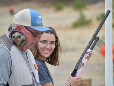 Youth participant in shooting sports holding pink firearm stands next to mentor. 