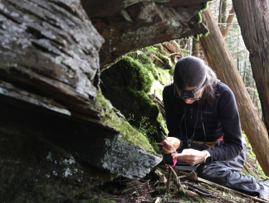Woman sitting on the forest floor examining something in a small tray she is holding