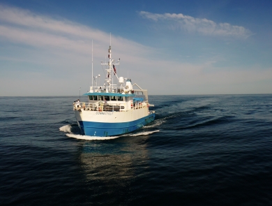 The Research Vessel Connecticut, a 90' steel blue and white ship with a tall mast and various antennas, is traveling through the water with no land in sight. The horizon stretches out far in the distance. The sky is blue, with white clouds.