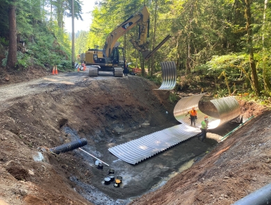 A construction site in the forest with a deep dirt pit with corrugated sheeting and workers wearing safety vests and hard hats. Heavy equipment and trees in the background.