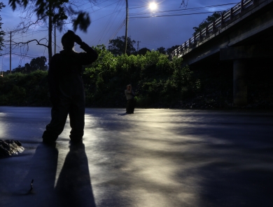 Two people standing in a river at night, looking up at a bridge