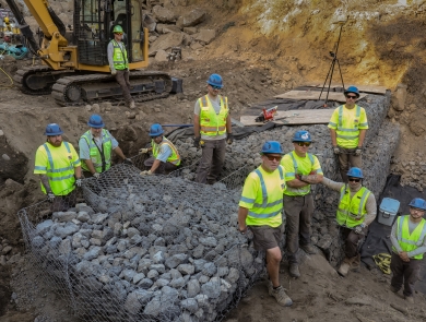 Ten people wearing bring yellow shirts and blue hard hats pose near a newly constructed rock wall
