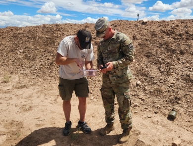a fish and wildlife service biologist and military personnel dressed in fatigues looks down at a container with a lizard