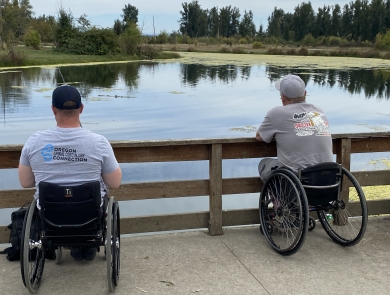 Two men in manual wheelchairs sit side by side fishing from a paved path, looking out over a calm pond bordered by trees and greenery, with a wooden railing in front of them.