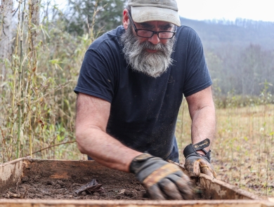 Man standing on the edge of a field, wearing work gloves, and sorting through dirt in a large sieve in front of him