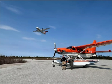 plane flying over two pilot biologists standing in front of a plane