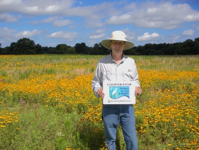 A person wearing a wide‑brimmed hat stands in a large field filled with yellow wildflowers. They hold a sign that reads “Cooperator – Partners for Fish & Wildlife – U.S. Fish & Wildlife Service.” Trees and a blue sky with scattered clouds appear in the background.