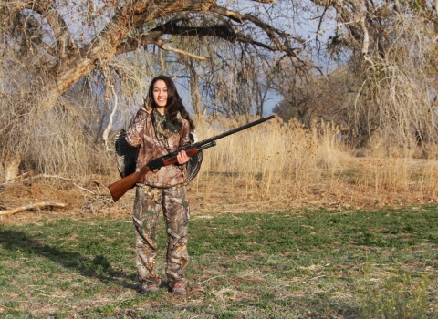 A young woman in camouflage holds a turkey over her shoulder and a gun across the front of her body. She is standing in a wooded area.