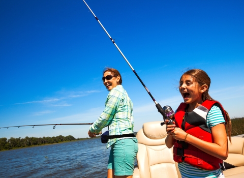 A young girl and her mother laugh as they enjoy fishing on a boat in the middle of a lake in the summer.