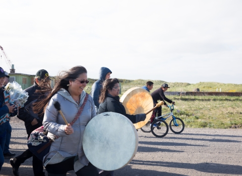 Woman with white skin drum leads group of people walking down a road. Kids on bikes flank her.