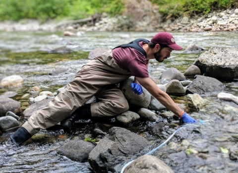 A man in waders, ballcap, and nitrile gloves holds an instrument in a river.