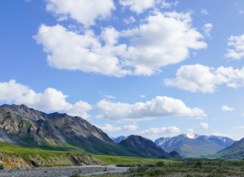 Landscape photo of green mountains, a river bank, and blue sky with clouds