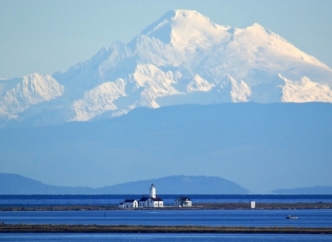 A distant view of a white lighthouse on a spit of land amid deep blue water with a snow-capped mountain looming in the background