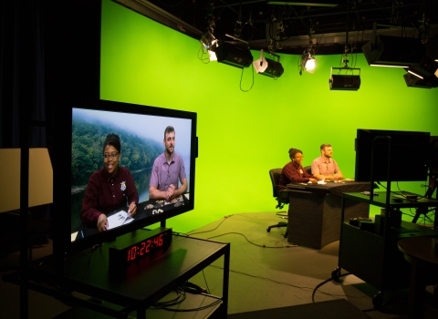 man and woman sitting at news desk with green screen in background of broadcasting studio