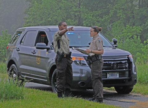 Two uniformed Refuge System law enforcement officers talk by a police vehicle and one points.