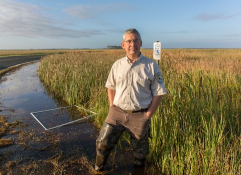 a man in Service uniform stands near a salt marsh with a refuge boundary sign behind him